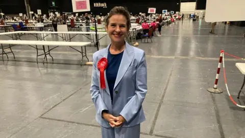 A woman stands in a hall filled with white tables. She is wearing a blue trouser suit and a red Labour party rosette.