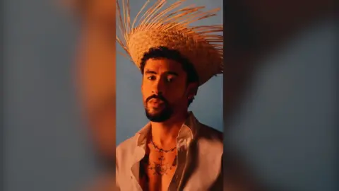 Bad Bunny has a black goatee and is wearing a straw hat and a cream shirt.