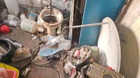 Specialist Cleaning Services A bathroom full of empty containers and waste, with a stained, damaged toilet in the middle