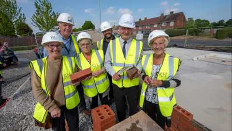 A group of six people wearing yellow high-visibility vests and standing in front of a block of bricks. Two women are holding a brick between them. There are houses and trees in the background. 