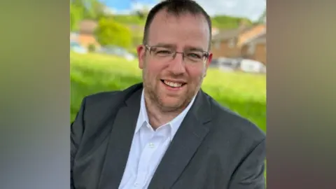 Matt Bishop/Labour Head and shoulders shot of Matt Bishop, Forest of Dean MP - he has short dark hair and is wearing glasses, a white shirt and a dark grey blazer. He is sat on a grassy bit of land on a housing estate, with houses and parked vehicles behind him, although the background is blurred.