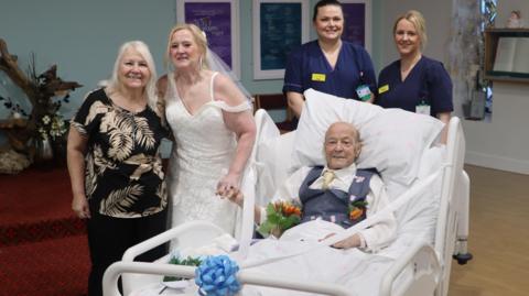 Malcolm Gretton in a hospital bed, with Maureen Draper in a wedding dress, along with family and staff