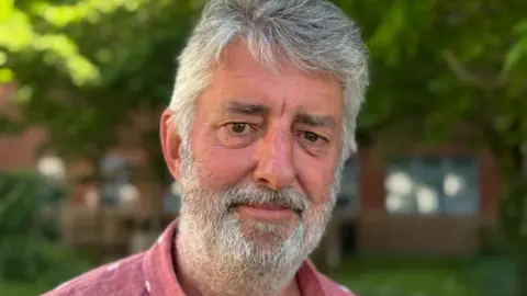 A man in a salmon pink shirt with grey hair and a grey beard. He is standing in a garden and smiling.