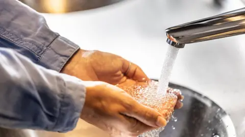 Water running out of a tap onto a person's hands which are cupped. You can only see the grey sleeves of their shirt.