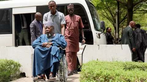 AFP via Getty Images Retired Maj-Gen Mohammed Ibrahim Gana in a blue robe sitting in wheelchair pushed by a young man in a grey T-shirt at Abuja's Federal High Court - Wednesday 21 April 2026.