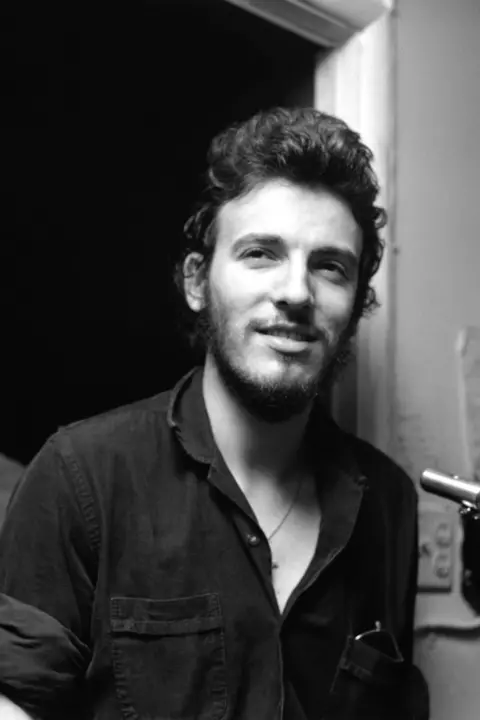 Getty Images Black and white photograph of Bruce Springsteen backstage at a concert, circa 1970. He has short curly hair and a close-cropped beard and moustache. Springsteen is wearing an open-necked black shirt.