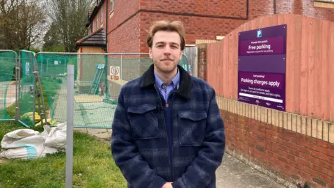 Luke is standing at the old pub site where the McDonald's could be built. He is wearing a checkered jacket and is smiling.