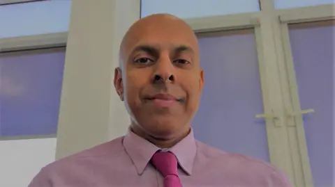 Supplied A man with a pink shirt and tie on, looking down at the camera. He is indoors.