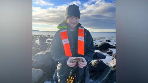 Nick Baker Horne Dr Nick Baker Horne is wearing a beanie hat and wax coat and orange scarf. He is standing in a coastal area holding a crab