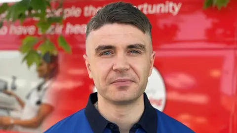 Stephen Huntley/BBC A man with short dark hair looks directly at the camera and smiles. He wears a blue and black rugby top. He is standing in front of a red van which is blurred slightly. 