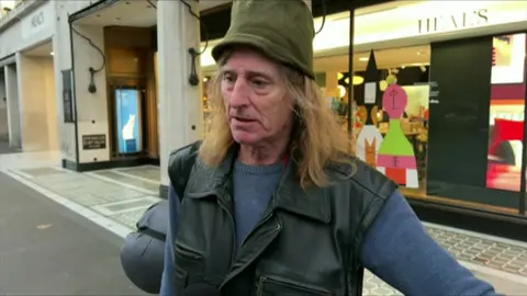 Gareth is seen in standing in front of a retail store in central London. He is wearing a green at, has shoulder length hair and is wearing a body warmer with his sleeping bag over his shoulder.