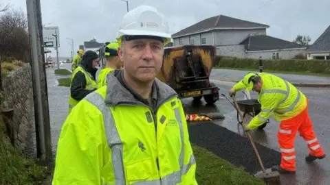 A man wearing a high viz jacket and a hard hat looks at the camera , behind him a team spread tarmac into a pothole as part of a repair.
