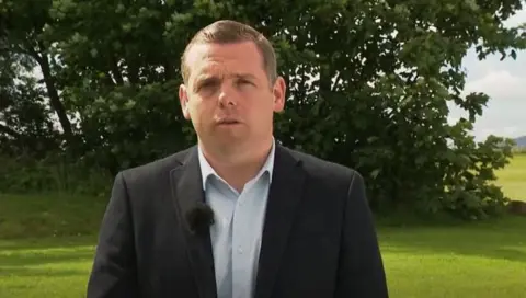 Douglas Ross wearing an open necked white shirt and black jacket, standing outdoors on grass with a large tree behind him