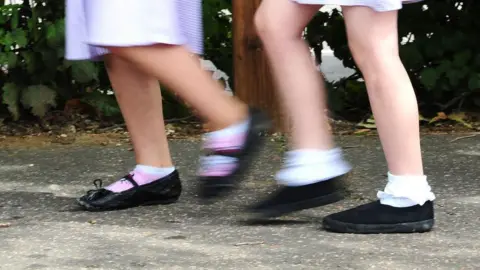 Two school girls feet moving along a pavement. They are both wearing black shoes and a summer dress. 