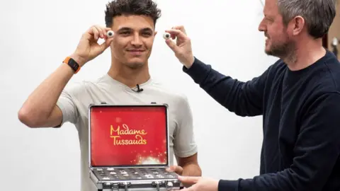 Madam Tussauds London Image of a man smiling at the camera and holding up a false eye to his face. In a case in front of him is a selection of false eyes. A second man is holding a false eye up to the man's face, on his right.