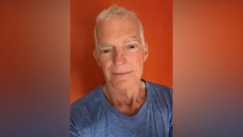 An older man with white hair. He is smiling at the camera and wearing a blue t-shirt.
