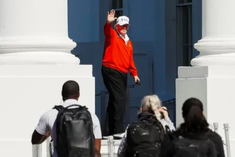 Aaron Schwartz / Reuters President Donald Trump, wearing a white USA cap and a red golfing sweater, waves as he passes by white pillars outside the White House