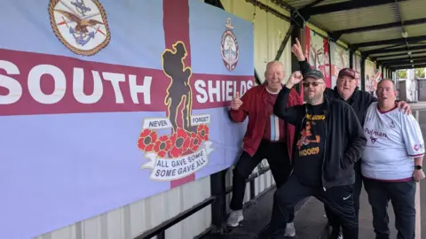 In a white, corrugated metal-backed stand, four supporters aged over 50 huddle together and raise fists in a joyous cheer. To their right hand side is a large South Shields flag in the clubs traditional claret and blue colours. 