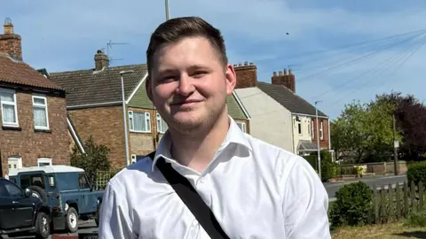 Frankie is wearing a white long‑sleeved shirt with a dark shoulder strap across the chest. He is standing on a residential street on a sunny day, facing the camera. Behind him are rows of brick houses with tiled roofs, parked vehicles along the road, a lamppost, low garden fencing, and patches of grass and flowers. Overhead wires and a blue sky with light cloud are visible, giving the scene a suburban neighbourhood setting.