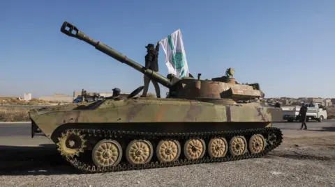 Reuters Seorang pejuang pemberontak Suriah mengibarkan bendera Hayat Tahrir al-Sham (HTS) di atas sebuah tank di Saraqeb, provinsi Idlib, Suriah (1 Desember 2024)