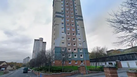 A tower block with blue and red patterns on the side of it. It is in a residential area with other houses and tower blocks in the background.