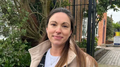 BBC A woman with her hair half up half down is wearing a beige coat and smiling at the camera