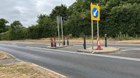 Clare Worden/BBC Image shows a two lane road with a traffic island in the middle which is where pedestrians should cross. There is an orange cone on the island to add to the visibility. 