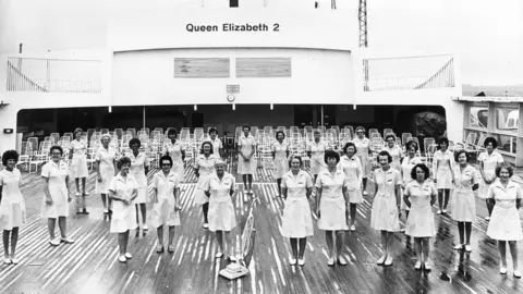 PA Media Dozens of women in their cleaning dresses pose with a vacuum cleaner on the deck of Queen Elizabeth 2 in 1975