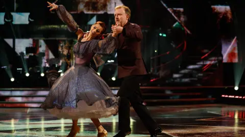 BBC/PA Chris McCausland and his Strictly Come Dancing partner Dianna Buswell dance on the set of the programme. Dianne stands in front of Chris with her back to him and her left hand raised whilst holding his right hand. She wears a dark purple dress and heels. Chris has his right hand on her shoulder and wears a brown shirt with black trousers and shoes.