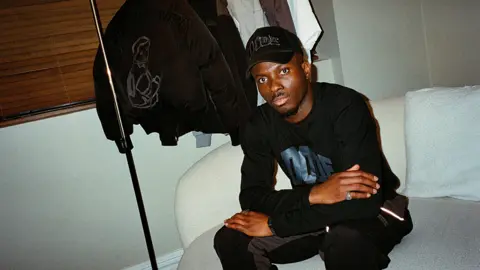 Dimeji Ode sitting on a sofa with a rack of his Dode clothing in the back. He wears a black baseball cap with the word "Dode" on it and wears a black top with "Dode" written on it in blue. His has black trousers with a grey vertical stripe. He looks into the camera and has a short goatee beard and moustache.