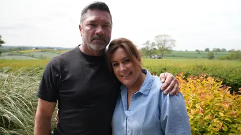 Matt and his wife pose for a picture, a field in the background. She has short, reddish hair which is tied up, and wears a light blue collared shirt.