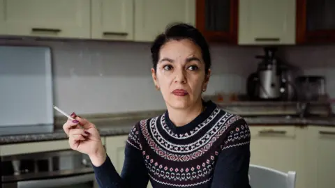 Enisa Mulic sits in her kitchen looking pensive while smoking a cigarette.