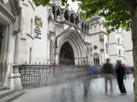 People walk by a large gothic building made from white stone, with the name Royal Courts of Justice written in large black letters.