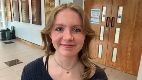 Kaitlyn us standing in a hallway with a door behind her. There are several brown boards on a wall with names of head girls and boys in gold. She was fair curly hair and is wearing a black jumper and a gold necklace. 