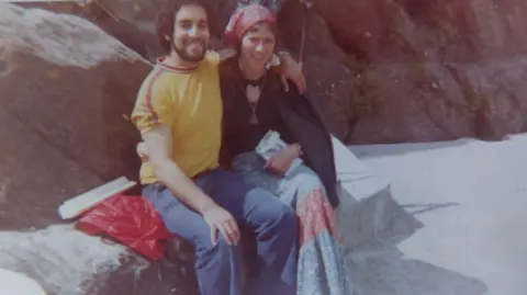 Supplied A man in a yellow T-shirt and jeans and a woman wearing a blue dress and black top sit on a rock, smiling towards the camera.