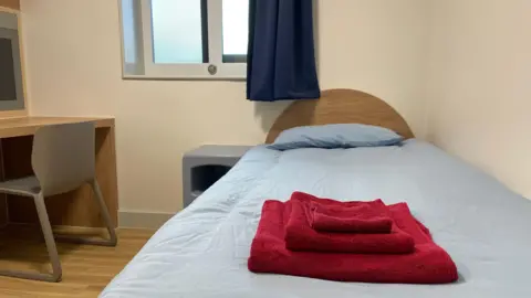 A single bed in a white room. The bed has a wooden headboard and a blue duvet. Bright red towels are folded on the bed. There is a desk and chair beside the bed and a window with a blue curtain behind it