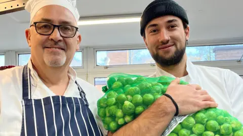 A smiling man in a chef's hat and apron stands beside a man holding a 9kg bag of sprouts