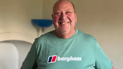 David is bald with a slight five o'clock shadow. He smiles at the camera while standing in what appears to be his house. He is wearing a turquoise Berghaus tshirt with the red and blue logo.