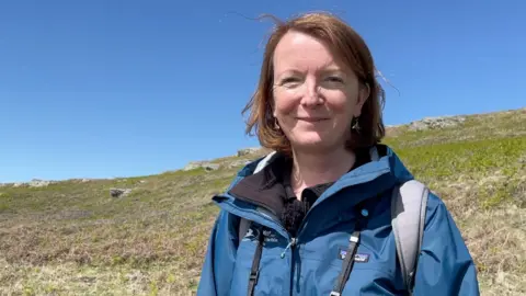 Lara Howe has short dark hair. She is wearing a blue outdoor jacket, with grey straps of a rucksack showing. She is standing in front of a grassy hilly and there are unbroken blue skies.