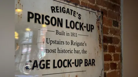 Zac Sherratt/BBC A sign in the Cage Wine Bar in Reigate, Surrey. The sign says: "Reigate's Prison Lock Up - Built in 1811". 