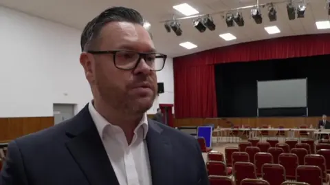 Craig Harris has dark hair and a short beard. He is wearing dark framed glasses, a white shirt and a blue suit jacket. He is standing in a hall where there are rows of red chairs and a projector screen in the background.