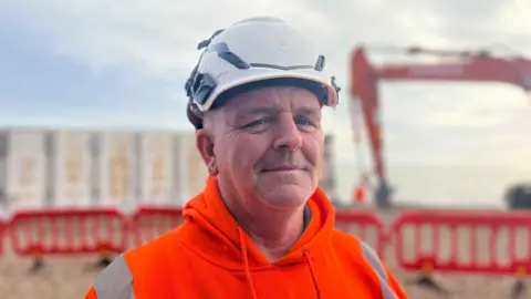 George Carden/BBC Worker Alan Macdowell wearing orange high vis and a white helmet with the container and an excavator behind him