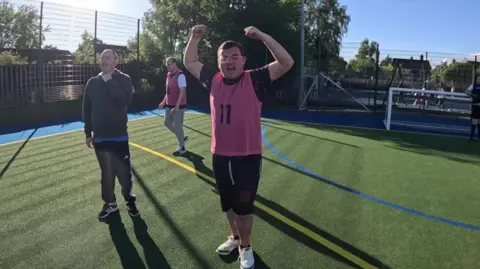 A person in a pink sports bib with the number “11” stands on a bright outdoor football pitch, raising both arms.
Two other participants walk nearby on the artificial turf, with a small goal and fence visible in the background.
The scene is sunny with long shadows, trees, and buildings surrounding the sports area.