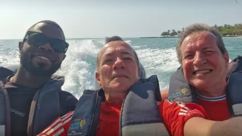 L-R Yostin Andres Mosquera, Albert Alfonso and Paul Longworth. They are shown smiling at a camera in a selfie, all wearing life jackets as they sit at the back of a speedboat. 