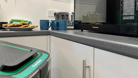 A grey and green bin labelled 'food waste' is in the foreground. Behind it there is a grey kitchen counter with cookies, bread and three mugs arranged in front of a toaster and next to a microwave.