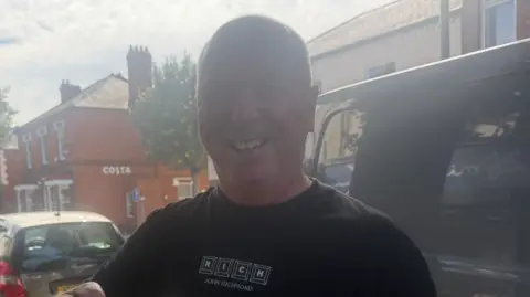 BBC / Sarah-May Buccieri The photo shows a man smiling at the camera. He's wearing a black t-shirt and is standing in front of a black van. There is a car and a row of buildings also behind him. 