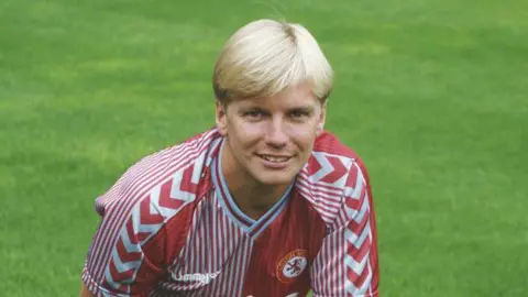 A man with short blonde hair in an Aston Villa kit, smiling at camera.  The kid is a mix of claret and blue stripes and arrows with the club's crest on his left breast. Behind him is green grass.