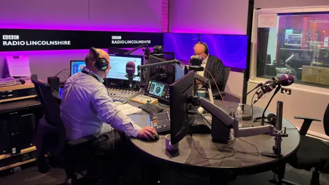 Beth Pritchard/ BBC Sean Dunderdale, a man wearing a light-coloured shirt sits behind a radio system. Facing him is Sir Edward Leigh, wearing a white shirt and a navy blazer. They are in a radio newsroom, surrounded by radio equipment.