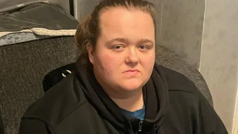 Anna sitting on a dark-coloured sofa looking directly towards the camera. She has light-brown tied-back hair and is wearing a black top