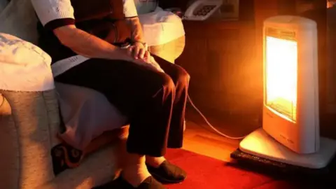 An elderly man, in his slippers and brown trousers, huddles by an electric fire